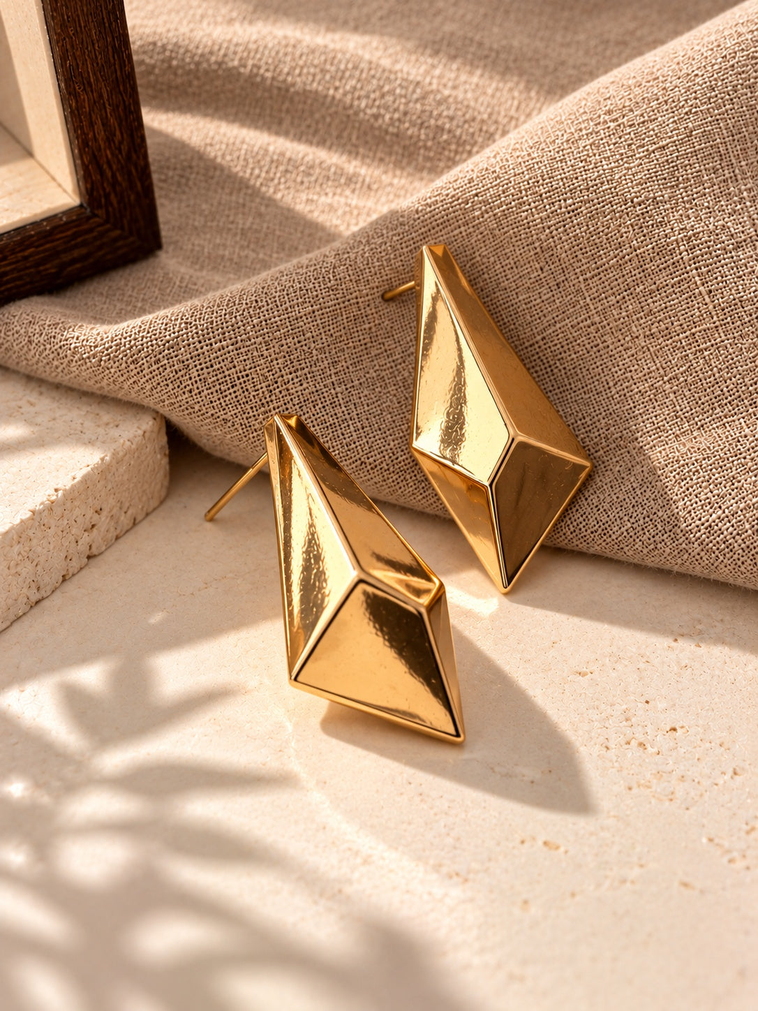 Gold geometric statement earrings on beige linen and stone backdrop, angle 4