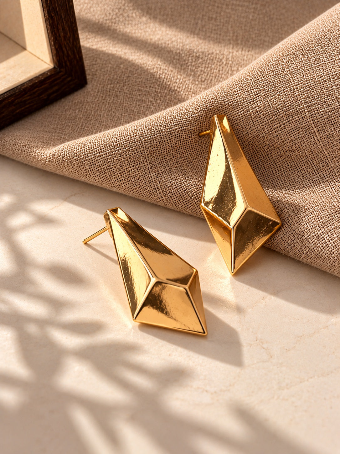 Gold geometric statement earrings on beige linen and stone backdrop, angle 3