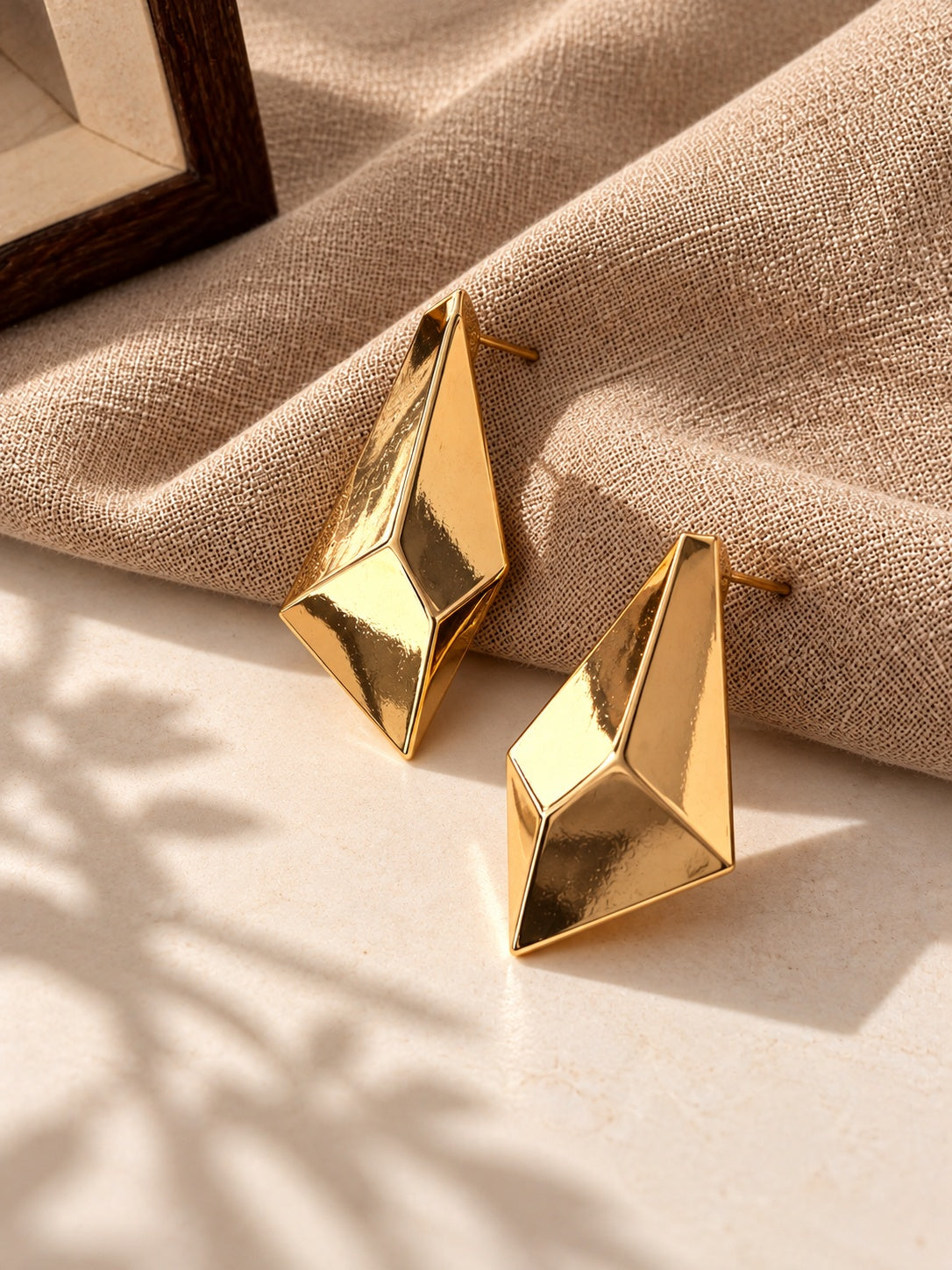 Gold geometric statement earrings on beige linen and stone backdrop, angle 2